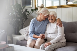 Two senior women sitting on a white couch, one embracing the other in a side hug, as they both smile together