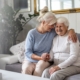 Two senior women sitting on a white couch, one embracing the other in a side hug, as they both smile together
