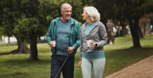 A senior couple in athletic wear walking their dog down a paved brick path, smiling at each other