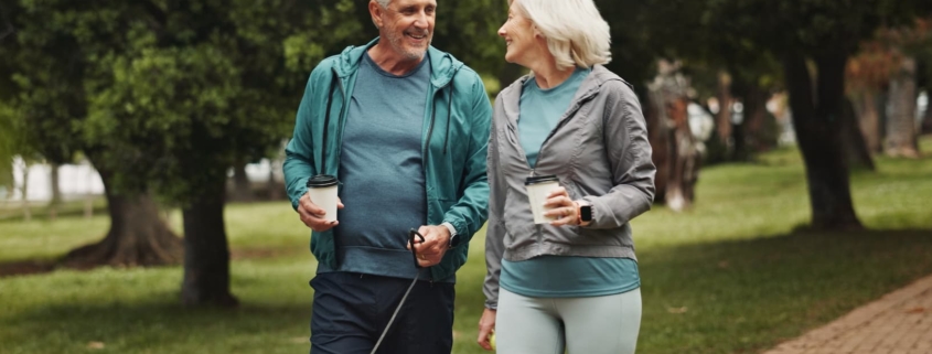 A senior couple in athletic wear walking their dog down a paved brick path, smiling at each other