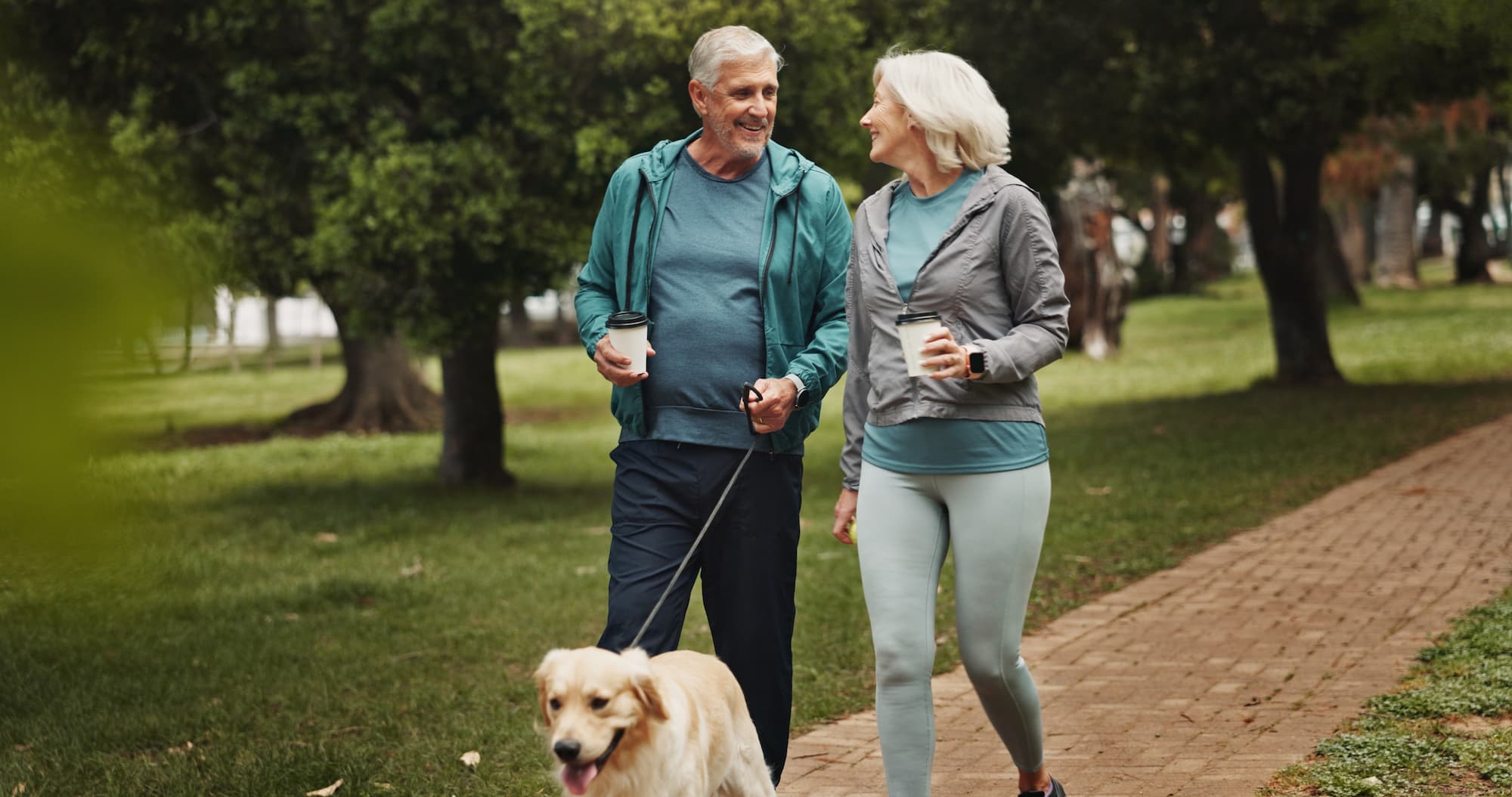 A senior couple in athletic wear walking their dog down a paved brick path, smiling at each other
