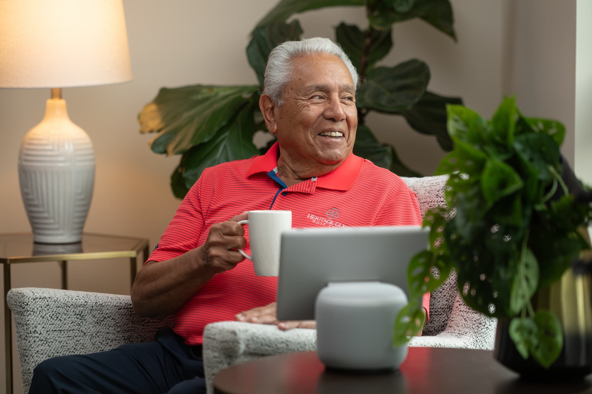 Resident of Heritage Community in Kalamazoo smiling as he drinks a cup of coffee as he sits on a chair