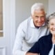 Elderly couple smiling as the man hugs his wife who is sitting in a wheelchair.