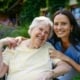 Senior woman in wheelchair and caregiver outdoors smiling.