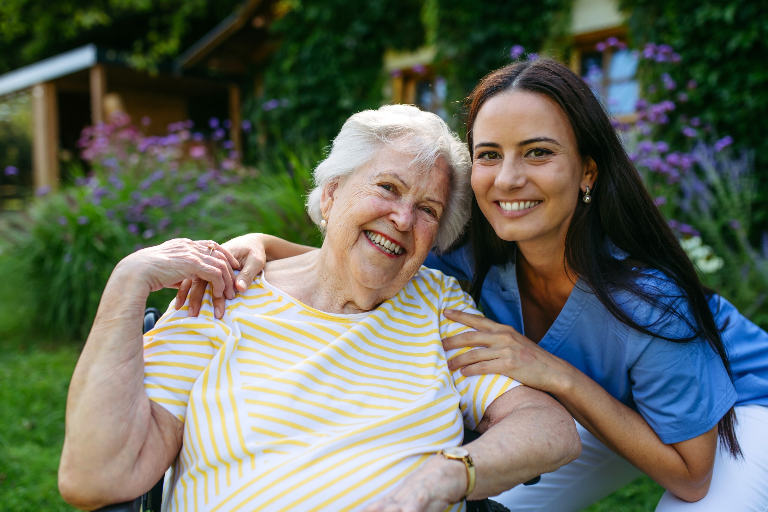 Senior woman in wheelchair and caregiver outdoors smiling.